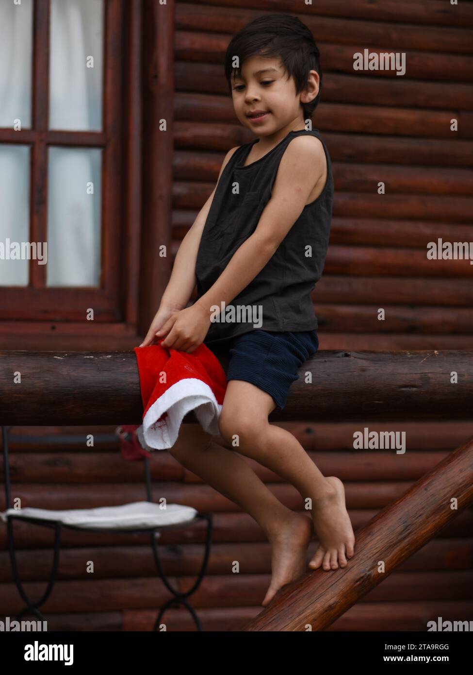 Garçon latin assis sur la balustrade en bois d'un balcon de cabine, tenant un chapeau de Noël, portant des vêtements d'été. vertical Banque D'Images