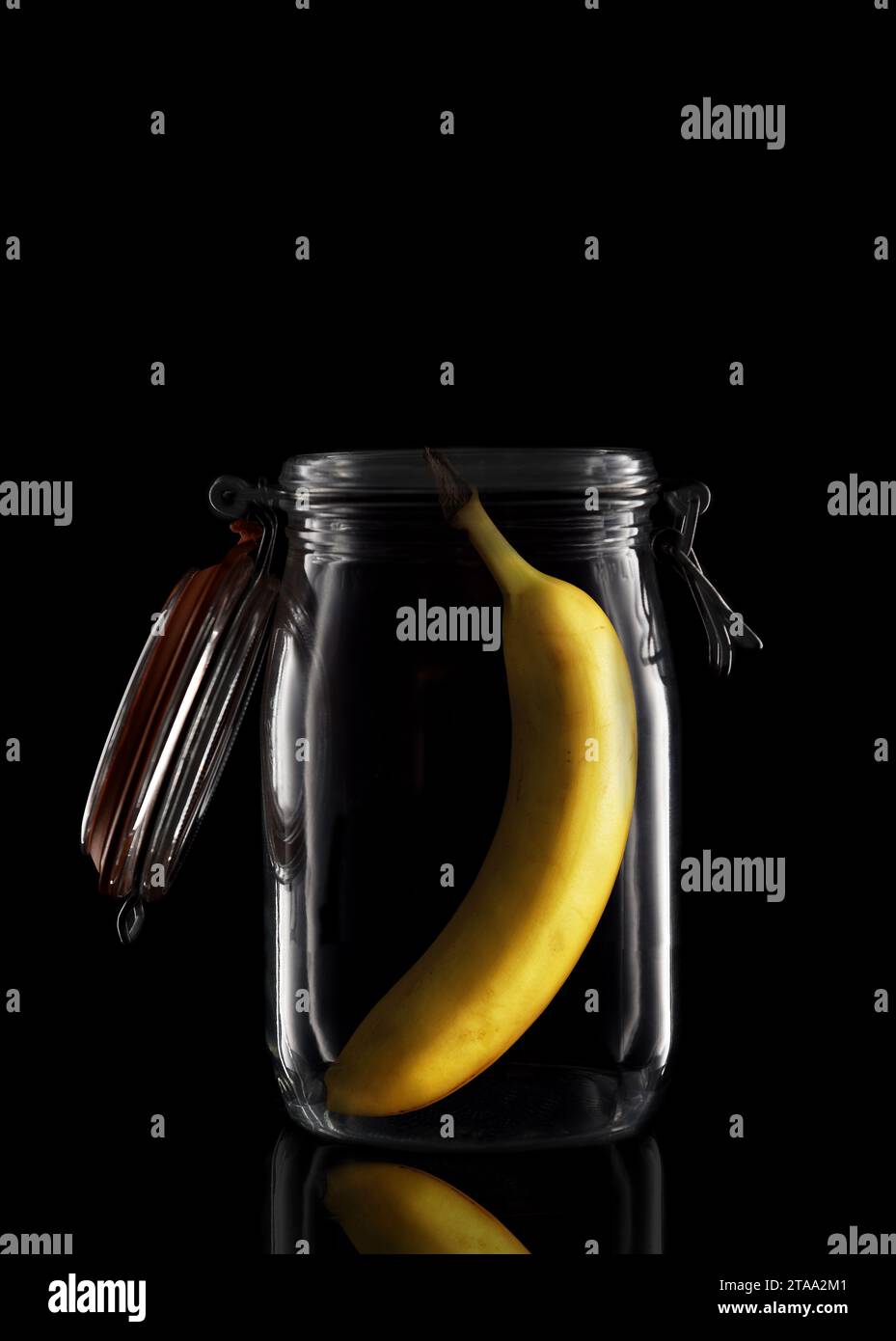 Une banane à l'intérieur d'un pot de stockage ou de conserve en verre isolé sur noir avec réflexion, avec couvercle ouvert. Banque D'Images