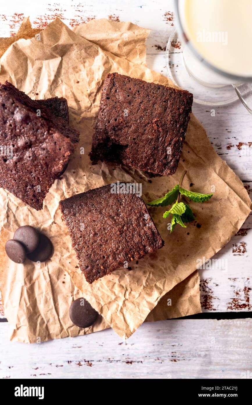 Beaucoup de morceaux de brownie au chocolat frais saupoudrés de cacao sur un papier de boulangerie avec un verre de lait sur la table blanche. Délicieuse tarte au chocolat maison Banque D'Images