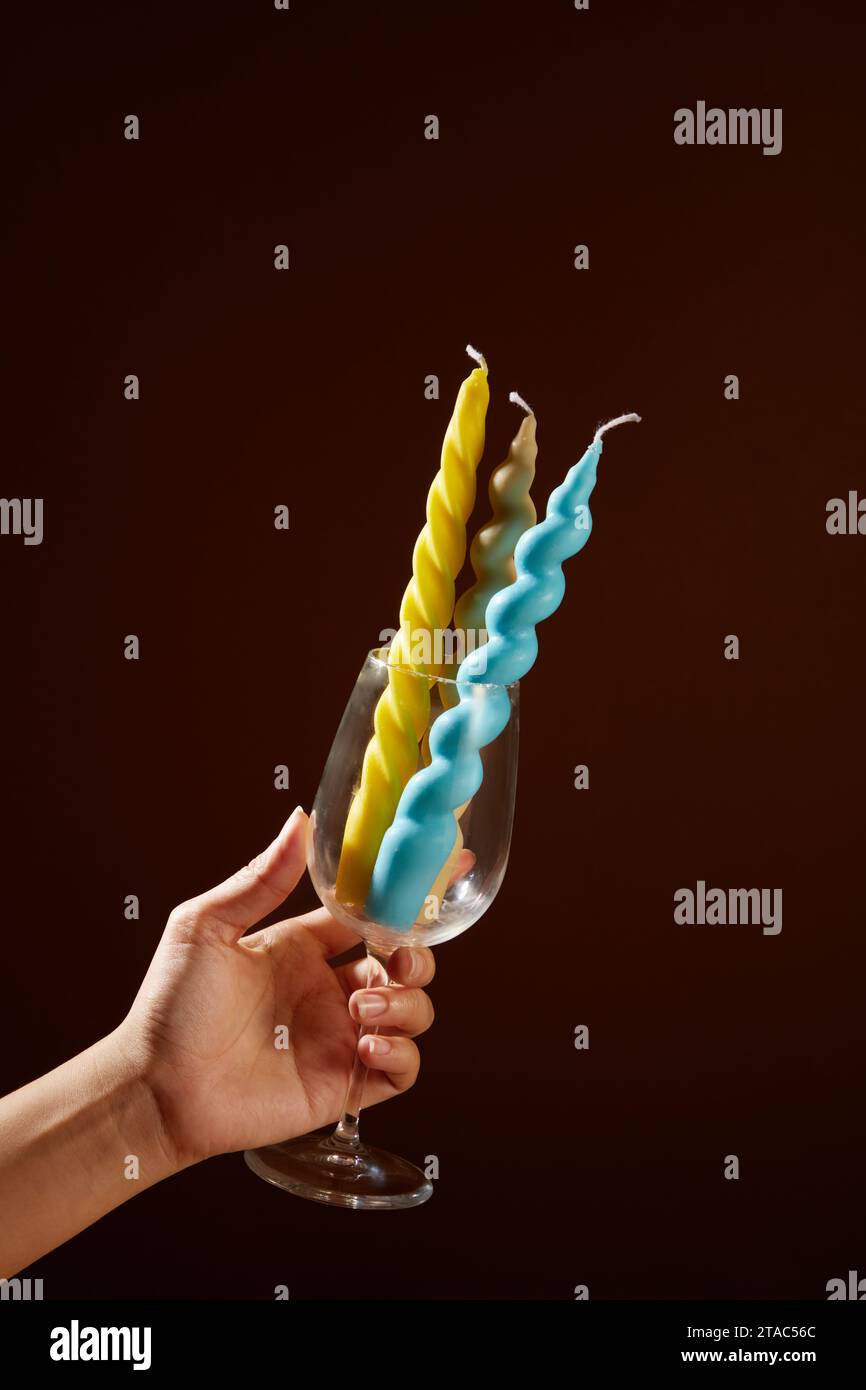 Une main tenant un verre à vin rempli de bougies colorées sur un fond brun foncé. Les bougies peuvent être fabriquées à partir de cire d'abeille, soja, quelques cires végétales, Banque D'Images