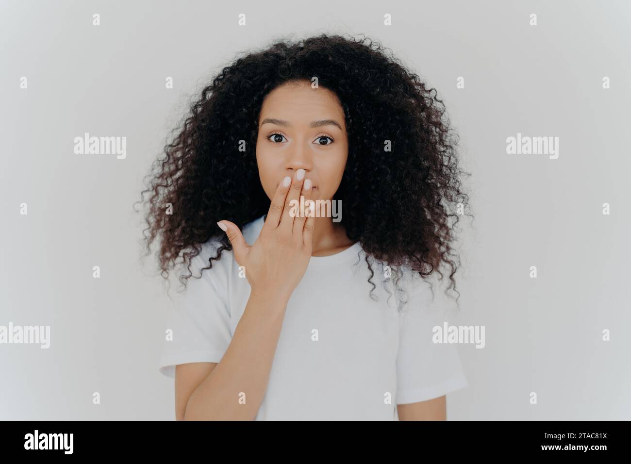 Femme noire surprise avec les cheveux bouclés, main sur la bouche, tee-shirt blanc, fond Uni Banque D'Images