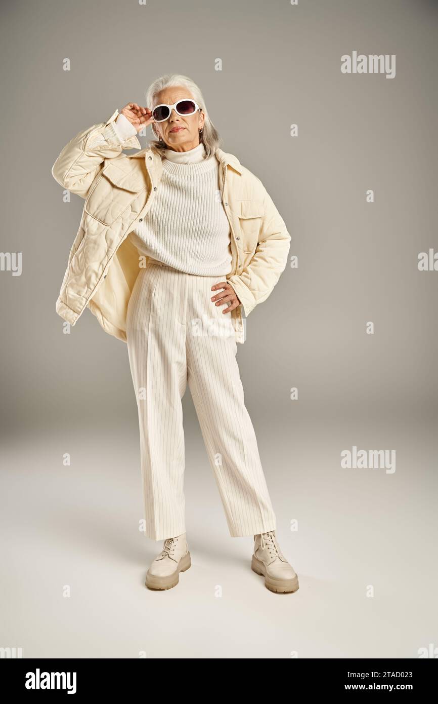jolie femme d'âge moyen en tenue d'hiver tenant des lunettes de soleil sur fond gris, main sur la hanche Banque D'Images