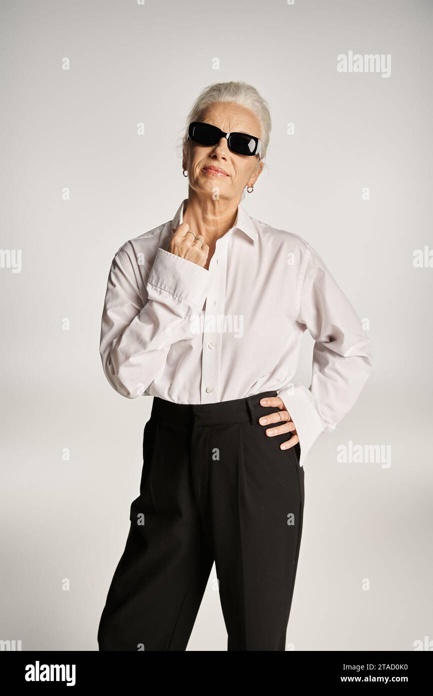 femme d'âge moyen à la mode en chemise blanche chic et lunettes de soleil posant avec la main sur la hanche sur le gris Banque D'Images