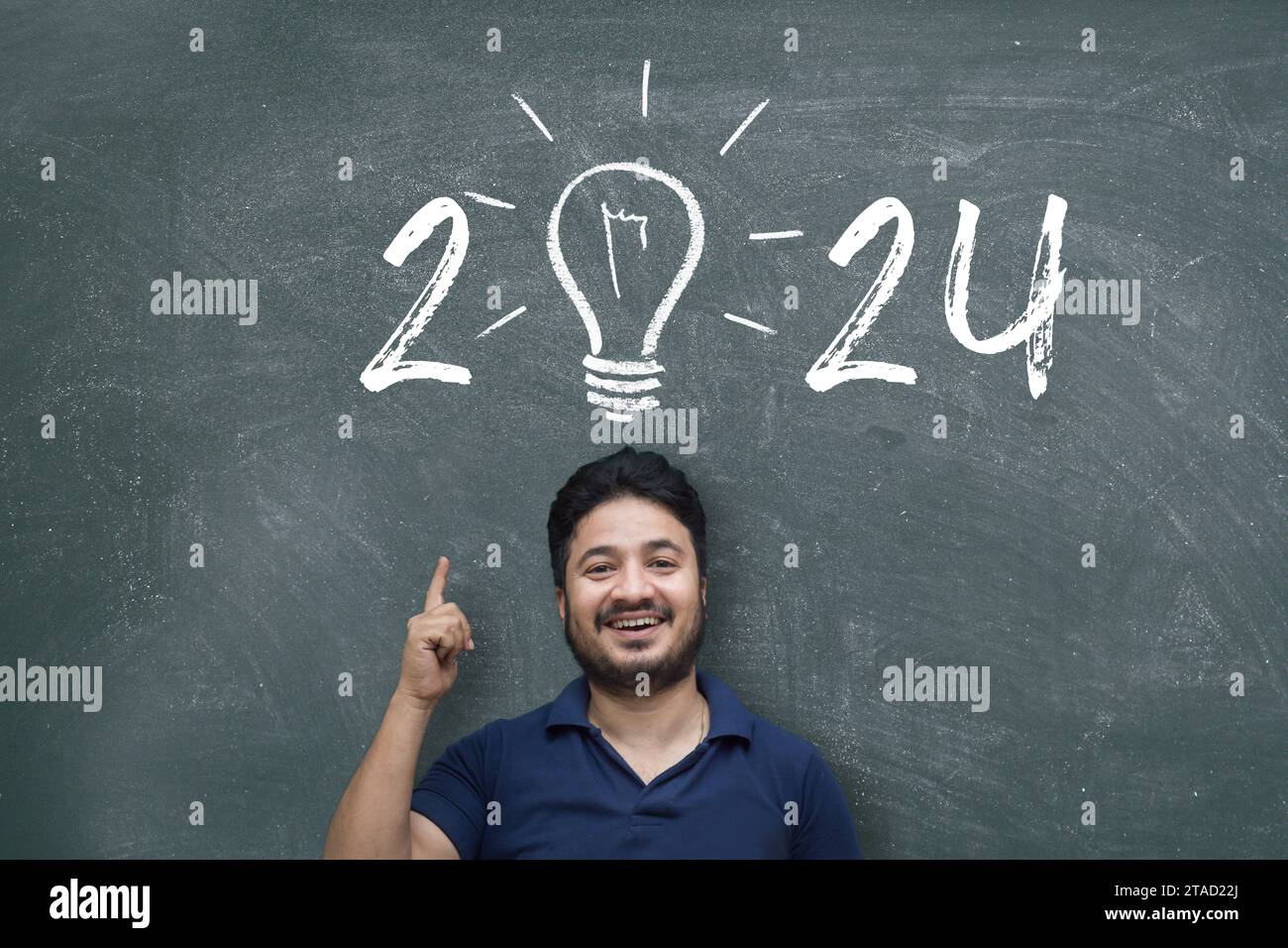 Indien / asiatique jeune homme pointant vers le haut tout en se tenant debout contre le tableau noir avec dessin à la craie de l'ampoule et nouvel an 2024 Banque D'Images