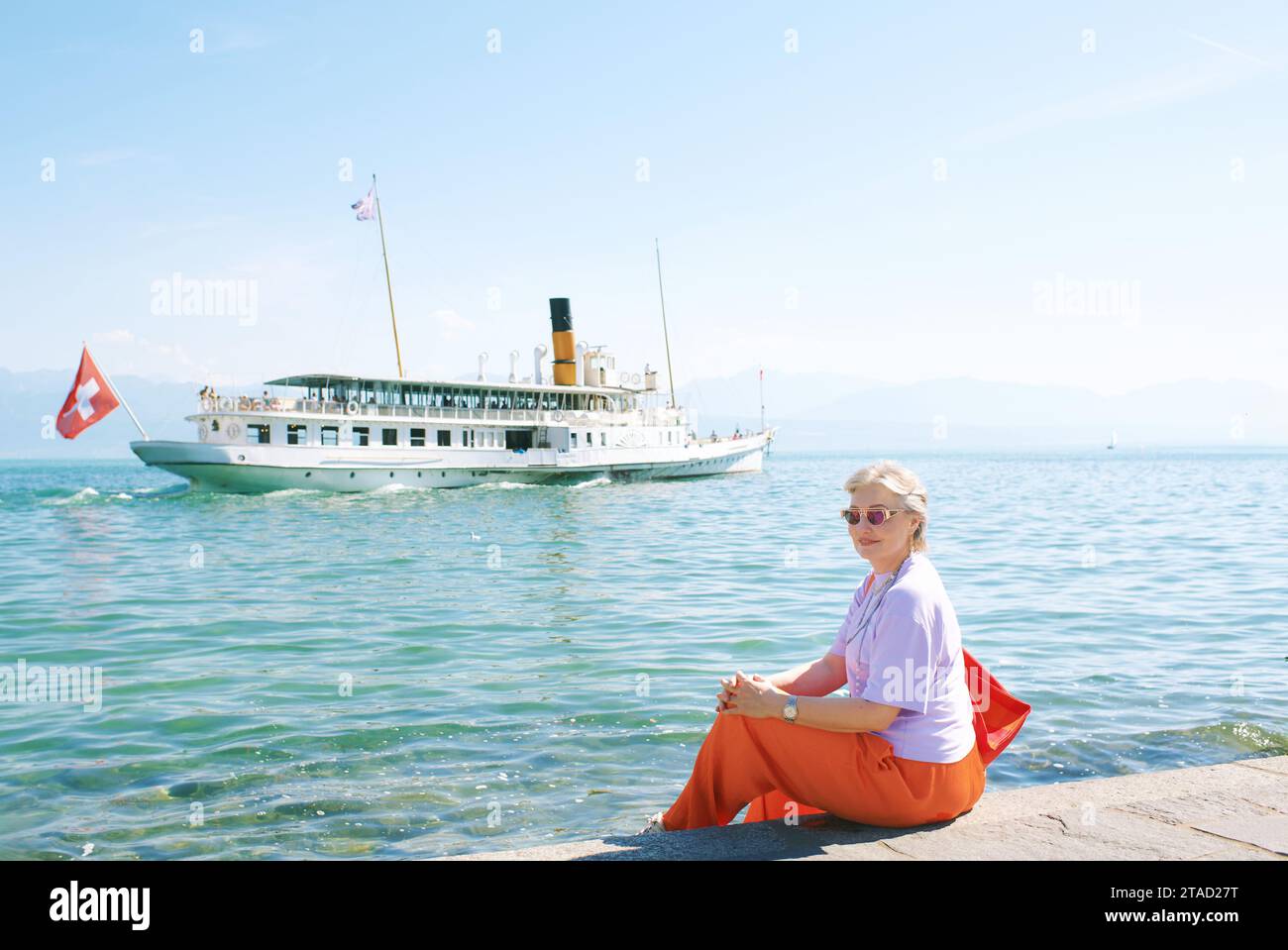 Portrait en plein air d'une femme âgée de 50 - 55 ans assise au bord du lac, regardant flotter bateau français suisse sur le lac Genève, Suisse Banque D'Images