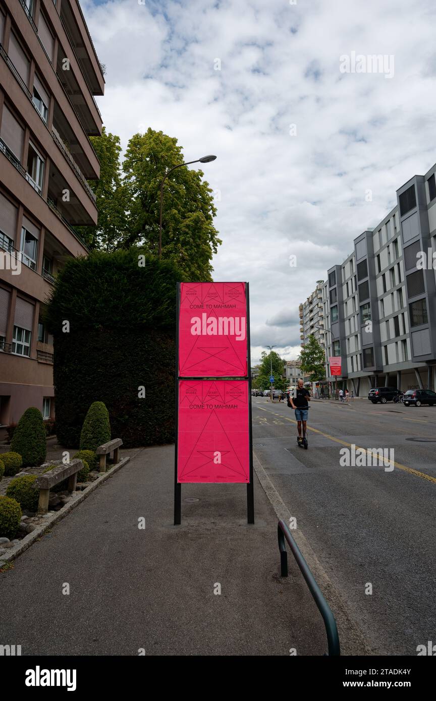 Une affiche rose vif annonce une exposition du musée à Genève, alors qu'un homme glisse sur son scooter, mêlant vie urbaine et art.. Banque D'Images