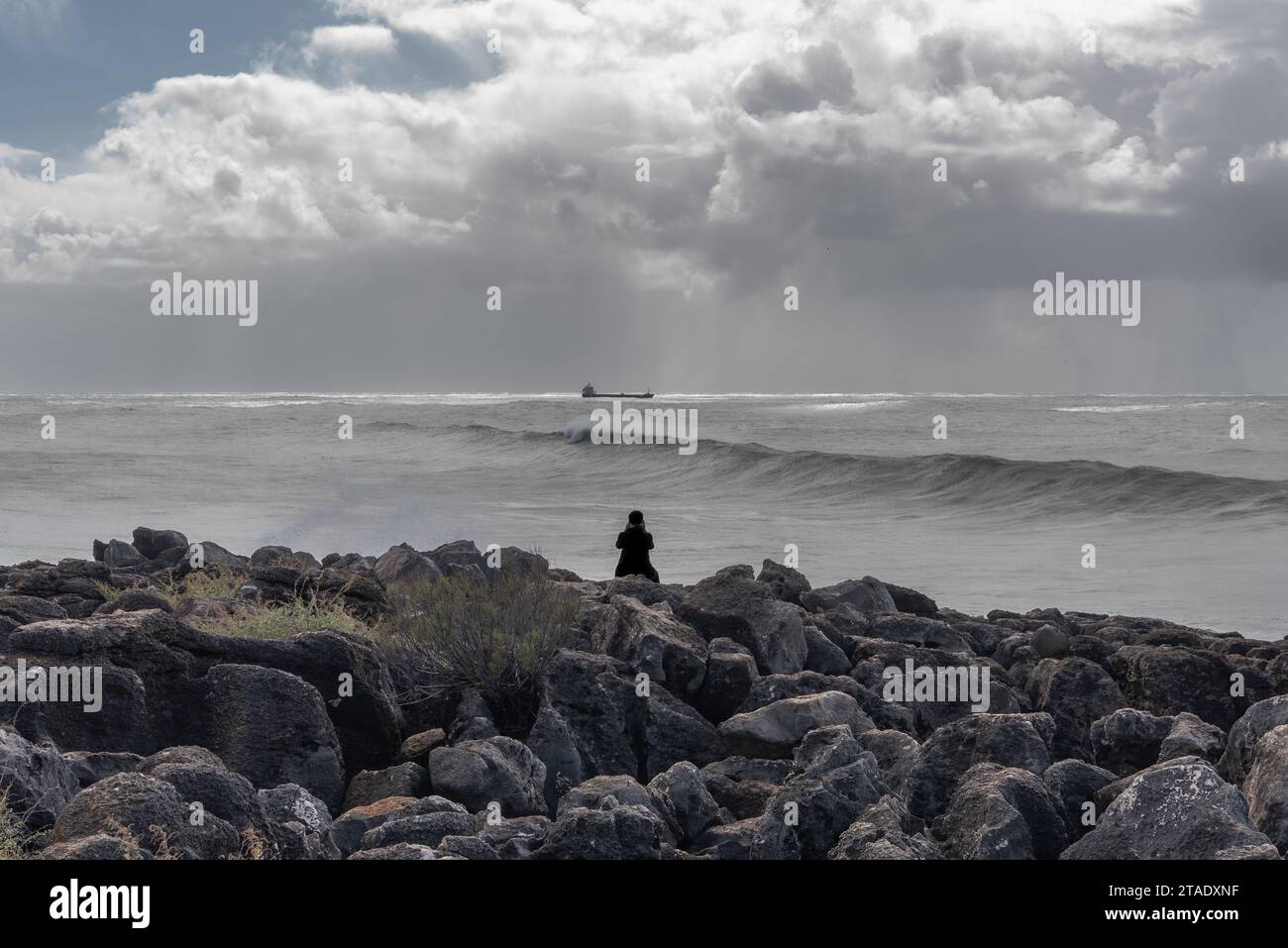 La silhouette d'une seule personne assise sur les rochers au bord de l'océan regardant un navire porte-conteneurs et de grandes vagues qui roulent dans l'océan Atlantique Banque D'Images