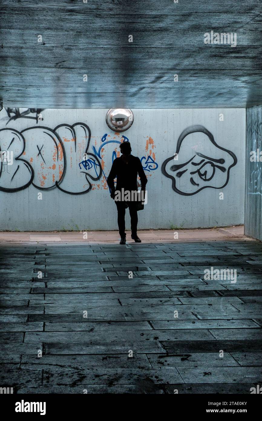 Silhouette d'une personne marchant dans un passage souterrain piétonnier sombre avec des graffitis sur le mur derrière lui. Banque D'Images