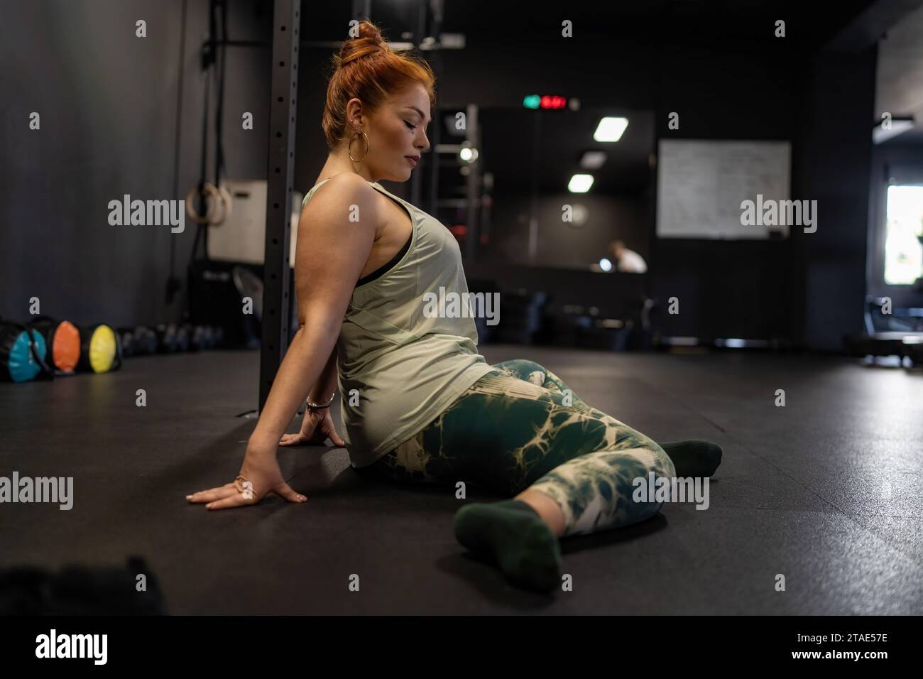 Une femme aux cheveux roux attachée dans un chignon, vêtue de vêtements de sport, effectue un exercice d'étirement sur le sol de la salle de gym. Elle est concentrée sur son entraînement, démonstrat Banque D'Images