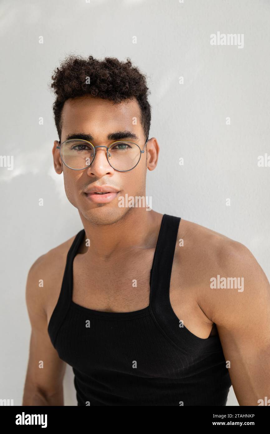 Portrait de jeune homme cubain bouclé dans des lunettes élégantes et t-shirt sans manches regardant la caméra Banque D'Images