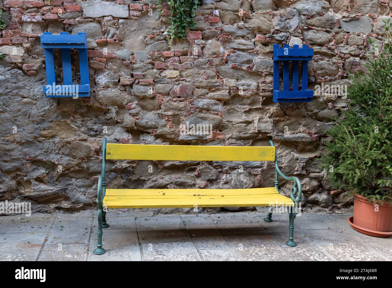 Un banc en bois jaune entre deux palettes en bois peintes en bleu et montées sur un mur de pierre et de briques vieillissantes à Sinalunga, Toscane, Italie. Banque D'Images