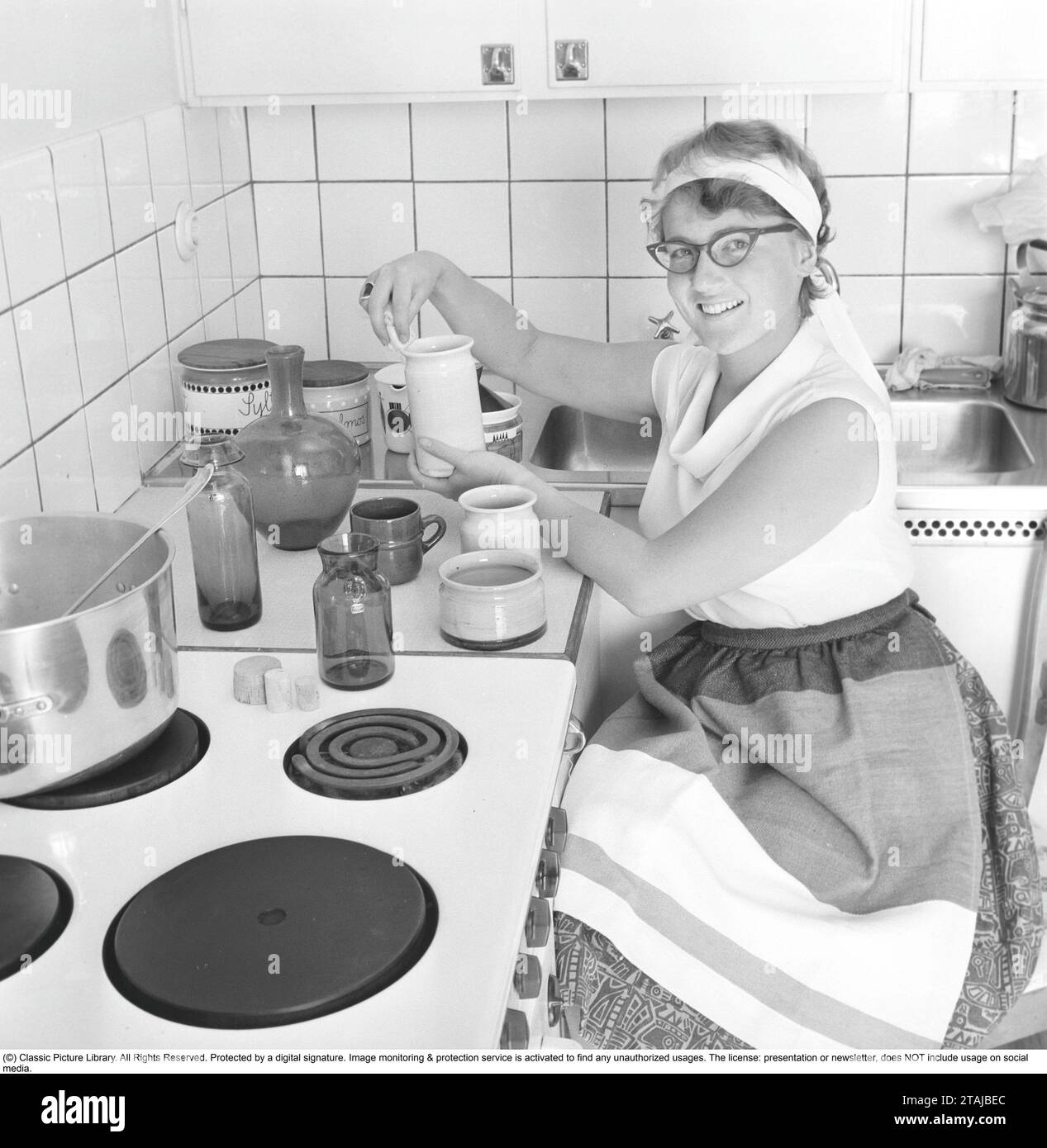 Dans la cuisine 1950s. Intérieur d'une cuisine et d'une jeune femme avec différents types de récipients, cruches, bouilloires en verre et pots. 1959 Banque D'Images