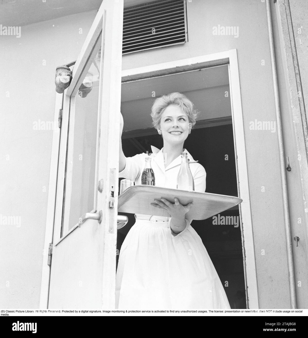 Une jeune femme travaille comme serveuse dans une pâtisserie et sort par la porte avec un plateau de boissons gazeuses. 1958 Banque D'Images