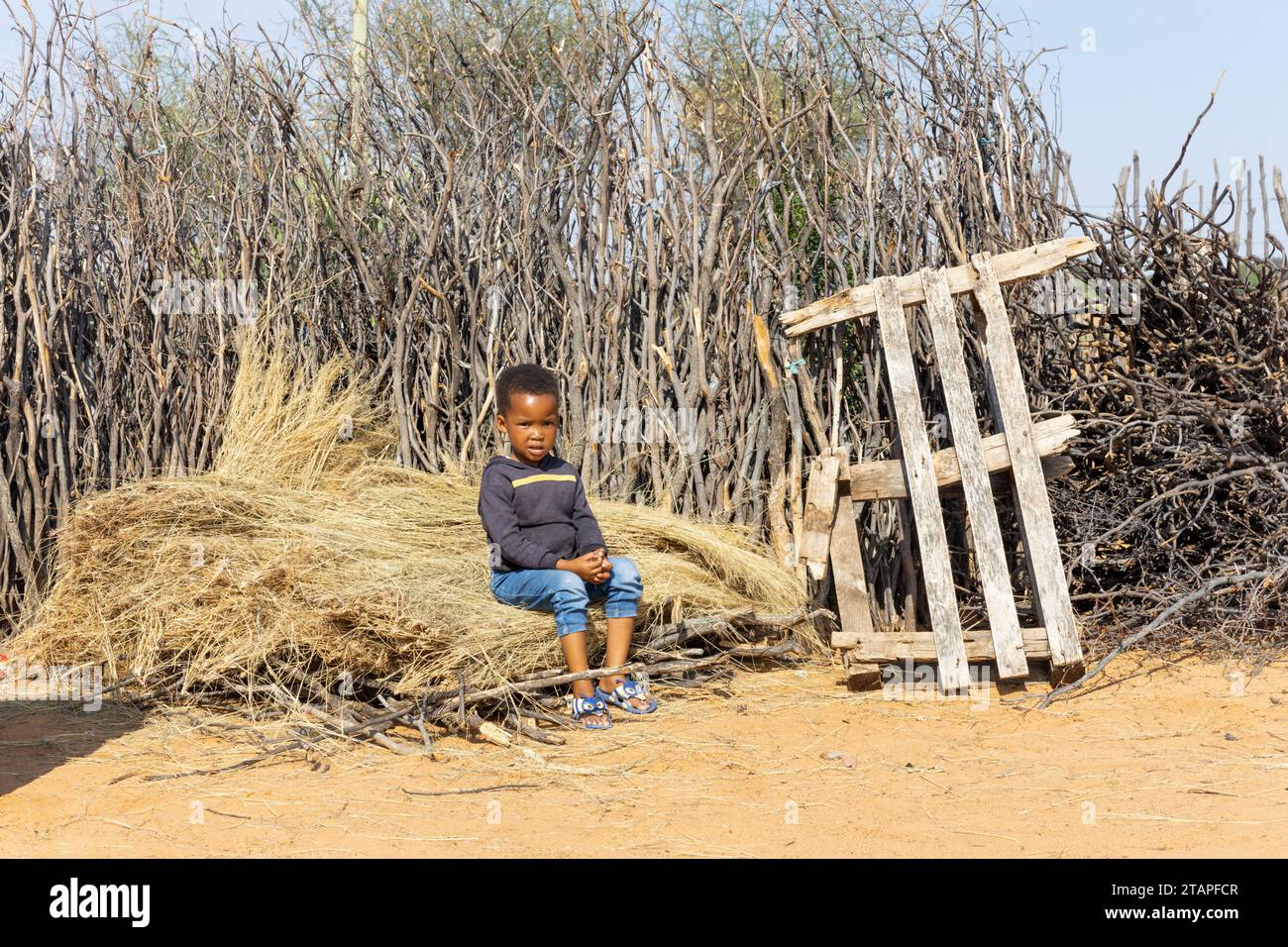 enfant de village africain situé sur un tas de chaume devant une clôture de branche dans la cour Banque D'Images