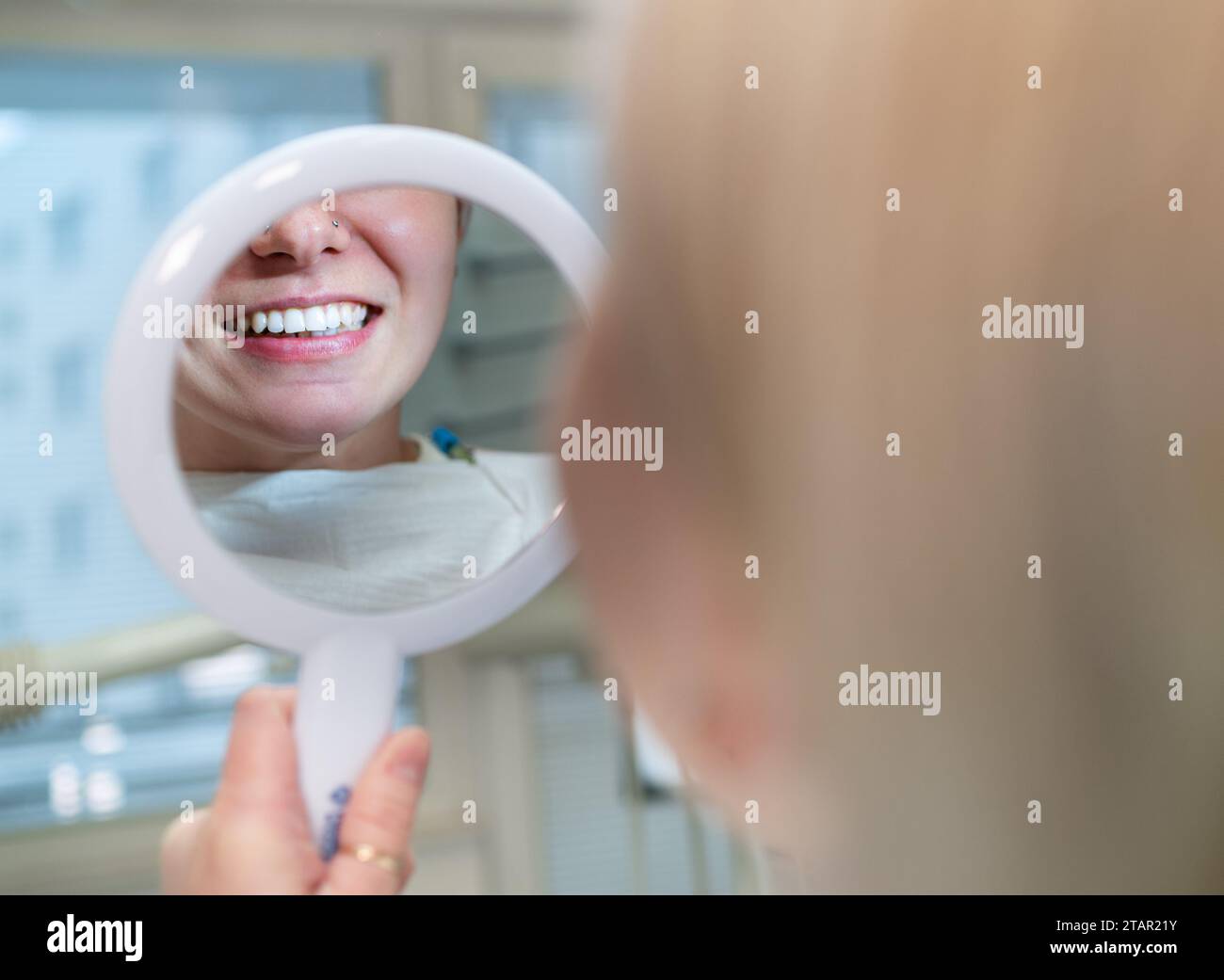 Jeune femme souriante assise dans une chaise de clinique de stomatologie et regardant le miroir évaluant le reflet de ses dents après la procédure de blanchiment dentaire. Santé c Banque D'Images