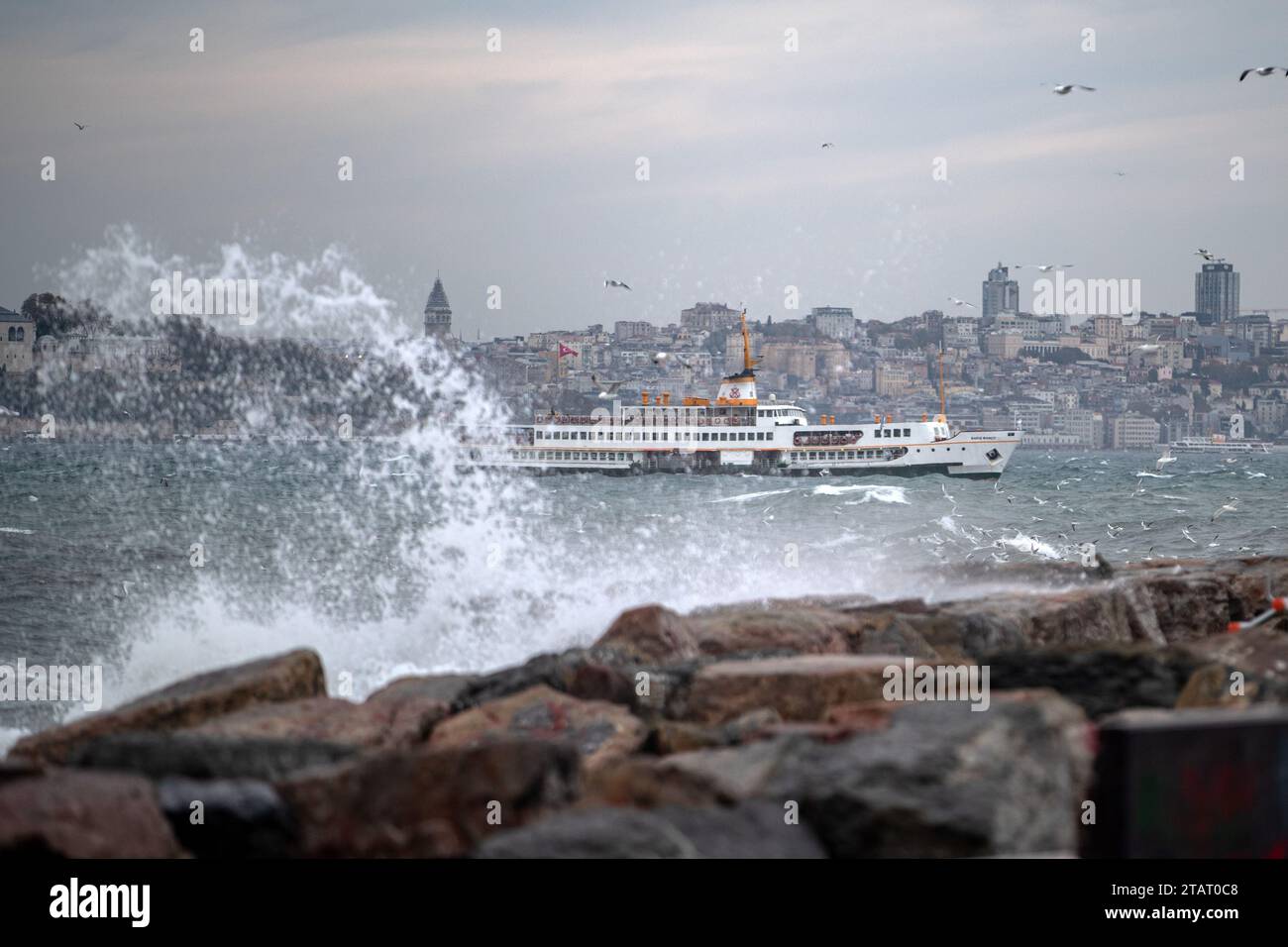 Un ferry des lignes de la ville vu passer derrière les vagues frappant les rochers à la plage de Moda. En raison du vent du sud-ouest affectant Istanbul, de hautes vagues ont été observées sur la côte de Moda. Banque D'Images