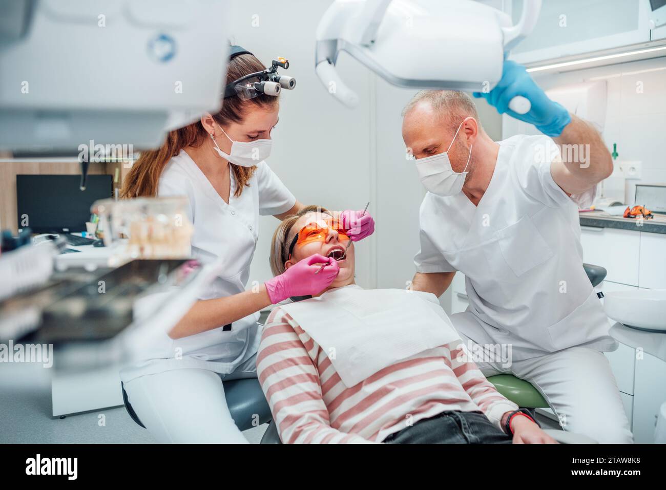 Rendez-vous en clinique dentaire. Femme dentiste en loupe faisant la prévention des dents à l'aide d'outils médicaux. Jeune homme assistant pointant la lumière. Banque D'Images