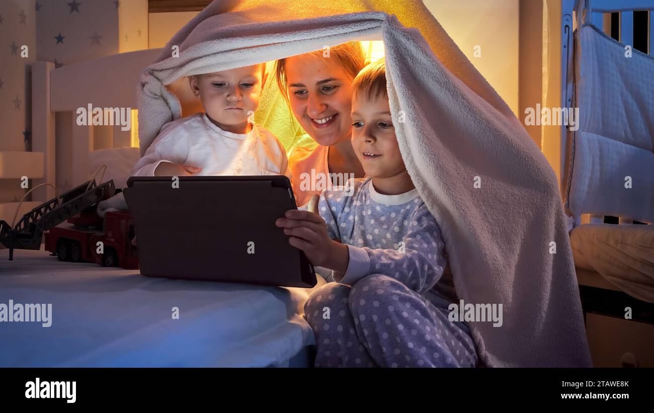 Portrait de deux garçons souriants en pyjama regardant la vidéo sur l'ordinateur tablette avec la mère la nuit. Famille ayant du temps ensemble, enfants avec des gadgets, pa Banque D'Images