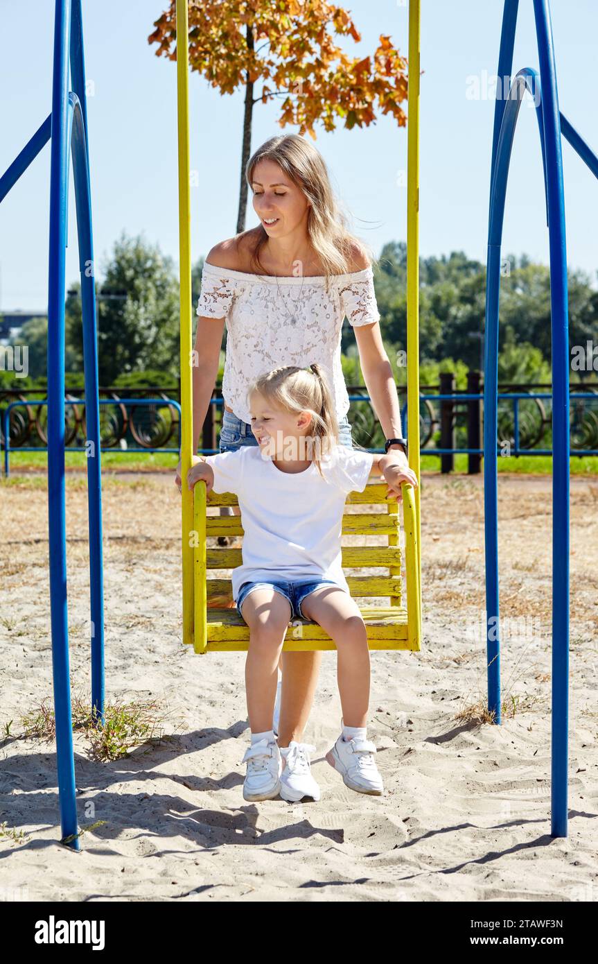 Mère avec fille sur balançoire sur une aire de jeux dans le parc de la ville d'été. Enfance, loisirs et concept de personnes - repos familial heureux et passer un bon moment Banque D'Images