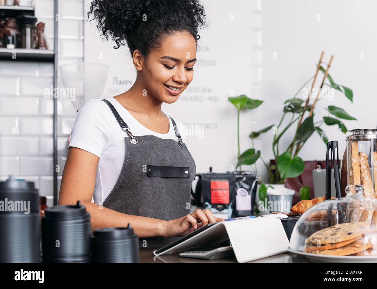 Barista en tablier tapant sur tablette numérique. Femme joyeuse avec les cheveux bouclés travaillant comme barista dans un café à l'aide d'une tablette numérique. Banque D'Images