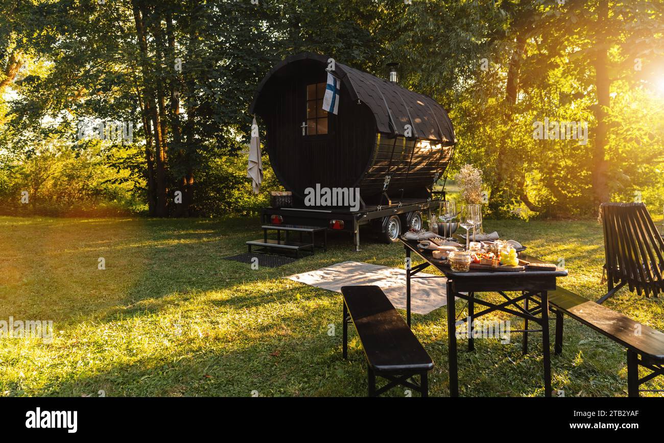Baril de sauna finlandais en bois avec une table avec des plats frais et des verres à vin dans un parc au coucher du soleil Banque D'Images