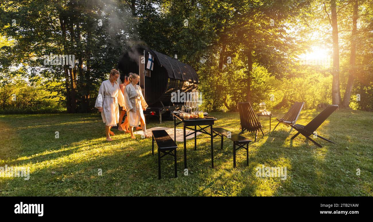 Les gens détendus en robes vient d'une cabine de sauna baril en bois dans un parc verdoyant luxuriant au coucher du soleil. Sont détendus, riant et profiter des vacances tout en se détendant Banque D'Images