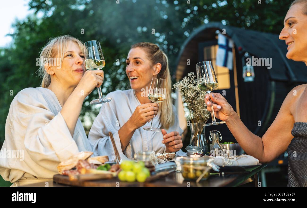 Femmes en peignoirs appréciant le vin en plein air, avec une cabine de sauna mobile finlandaise en arrière-plan Banque D'Images