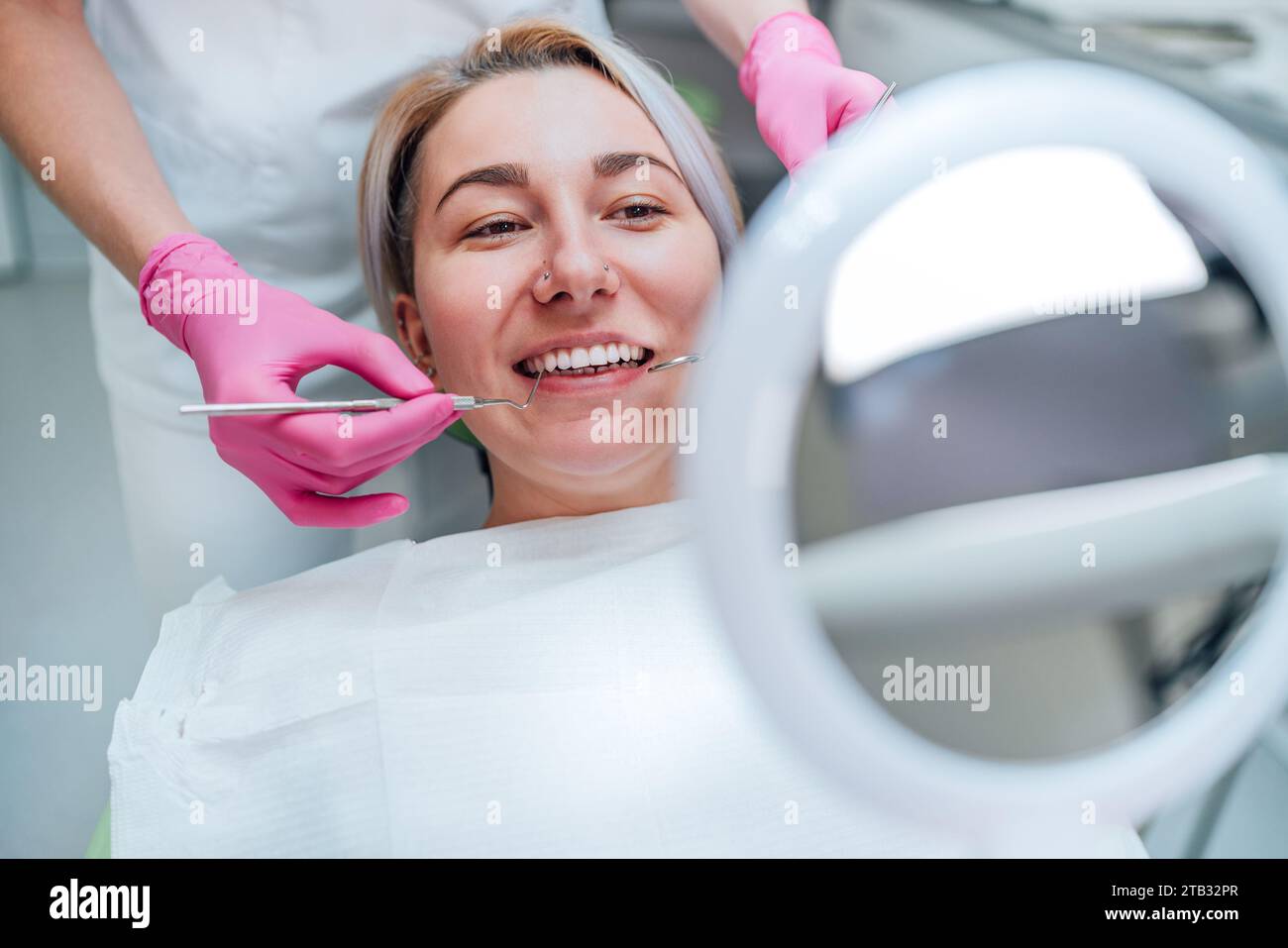 Portrait de femme souriante regardant le miroir dans la chaise de stomatologie et les paumes gracieuses du dentiste dans des gants roses avec excavatrice et miroir outils médicaux. Banque D'Images