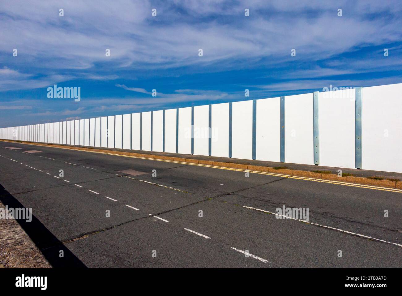Mur lambrissé de métal blanc avec une route goudronnée vide au premier plan et ciel bleu au-dessus. Banque D'Images