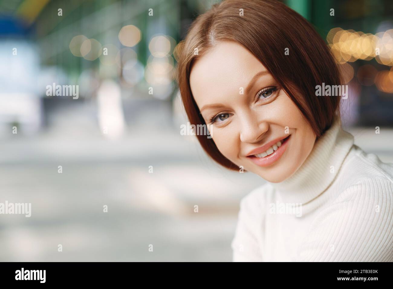 Femme souriante en col roulé blanc avec des lumières bokeh urbaines Banque D'Images