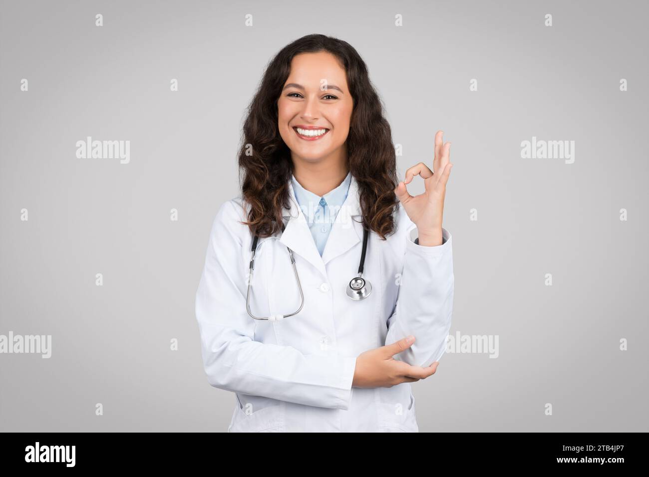 Femme médecin européenne heureuse en manteau blanc faisant signe ok avec la main, debout sur fond gris, souriant à la caméra Banque D'Images