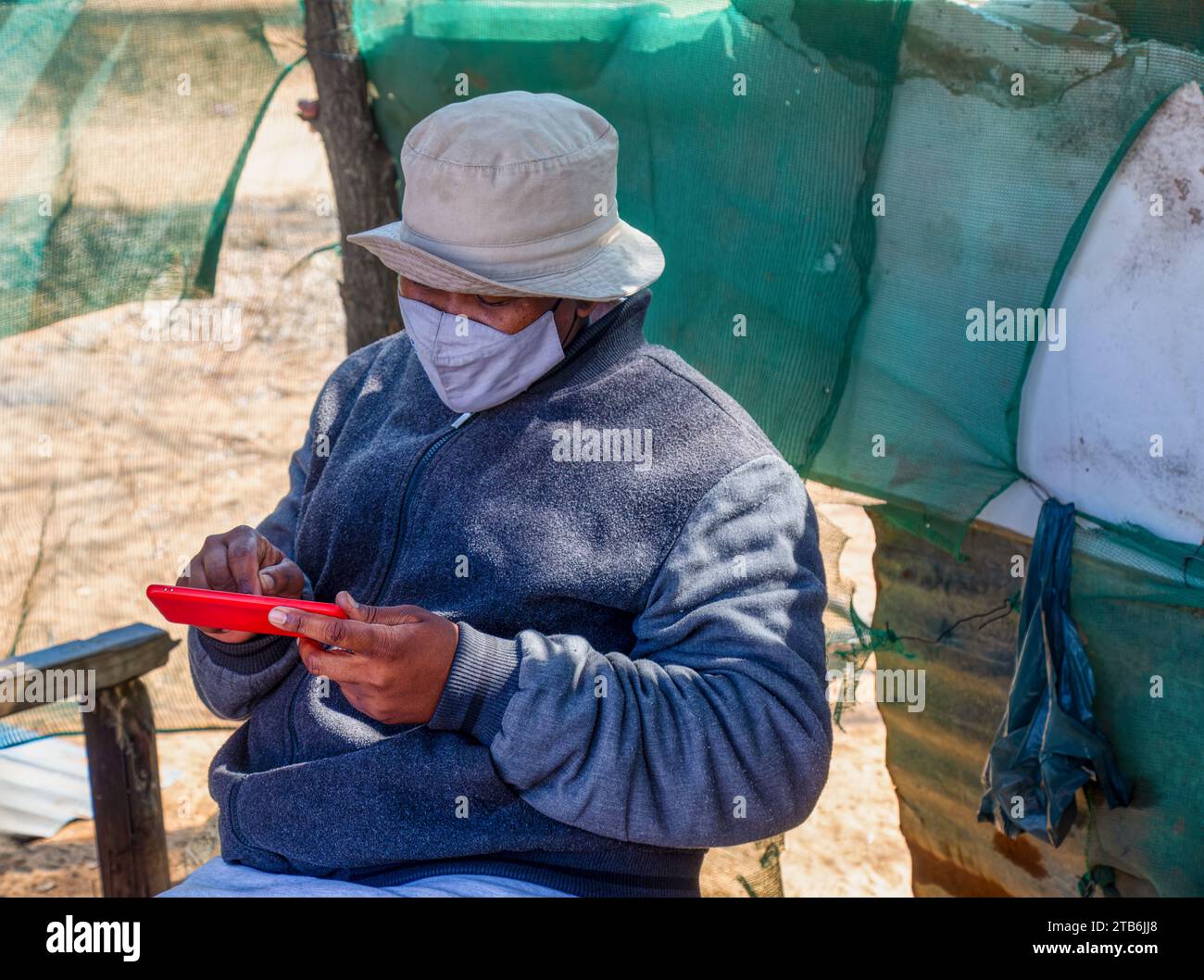 villageois , heureux en surpoids jeune homme africain portant un masque en utilisant le téléphone dans un hangar Banque D'Images