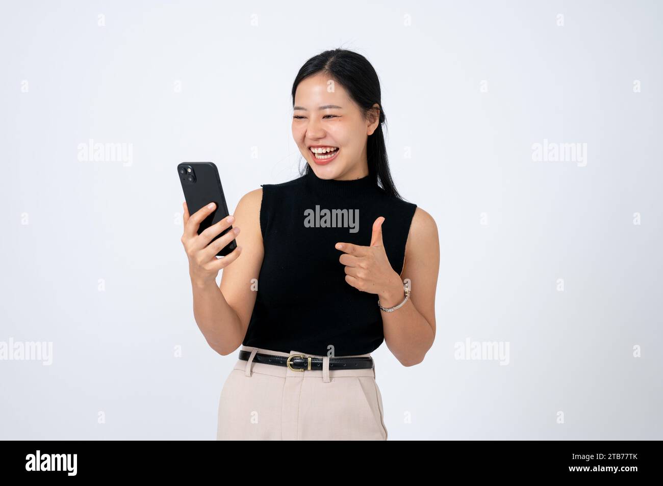 Une femme d'affaires asiatique joyeuse et belle pointe son doigt sur l'écran de son smartphone tout en l'utilisant, en parlant lors d'un appel vidéo, en regardant des clips o Banque D'Images