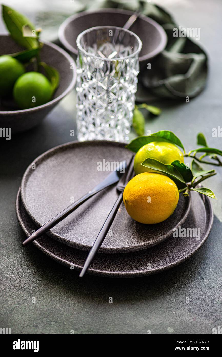 Un décor de table sophistiqué avec des couverts noirs sur une assiette sombre avec un citron jaune vif, un verre texturé et des citrons verts frais. Banque D'Images
