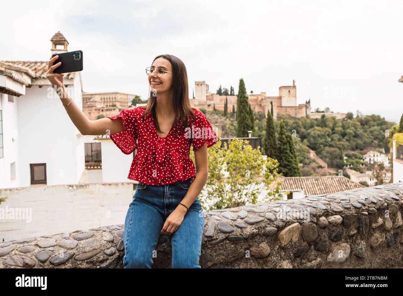 Jeune femme souriante habillée en casuals prenant selfie avec smartphone tout en étant assise sur le mur de soutènement avec Alcazaba forteresse en arrière-plan à Grenade, Banque D'Images