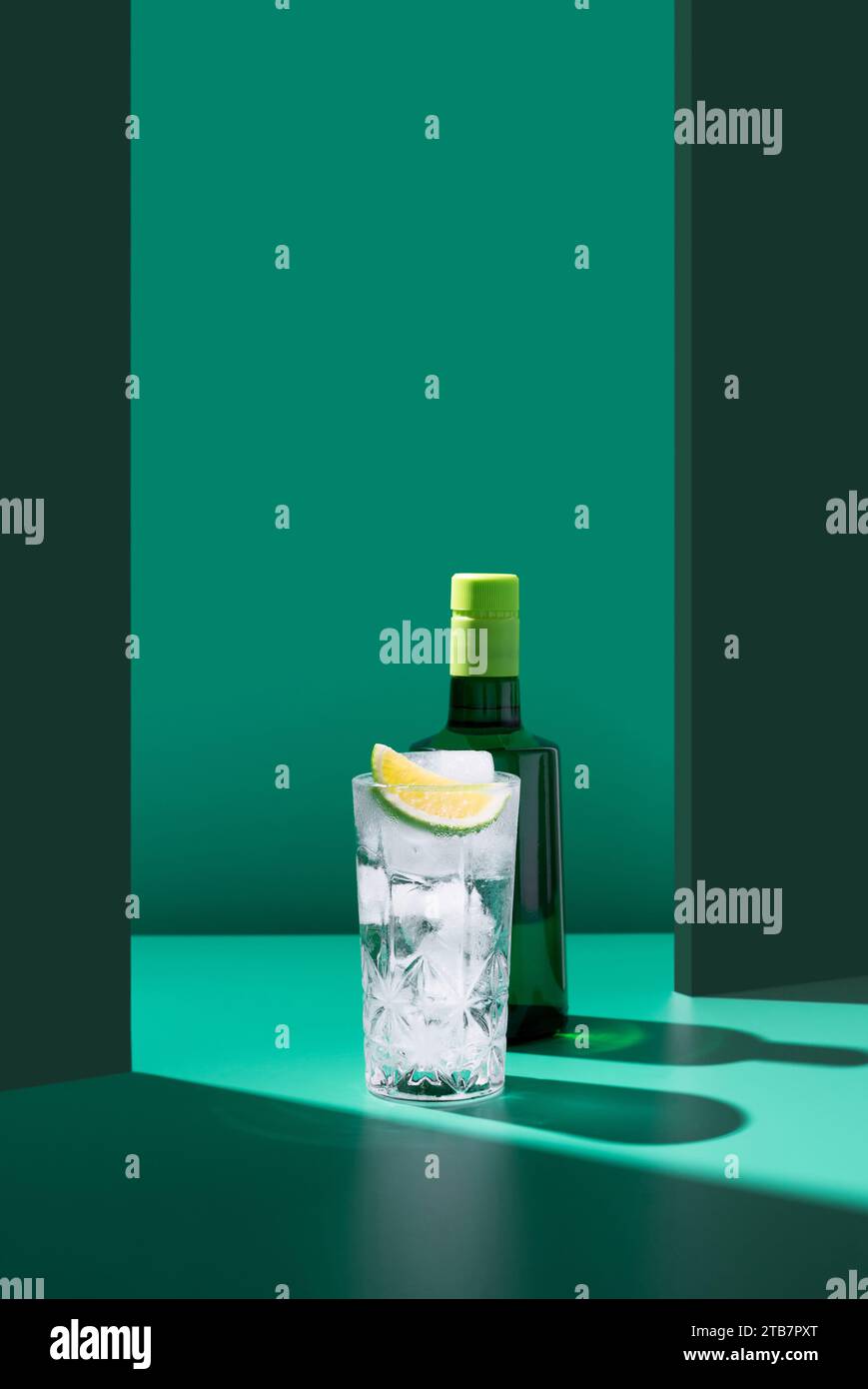 Une composition minimaliste avec une bouteille de gin derrière un verre d'eau tonique orné d'une tranche de citron vert, le tout sur un fond vert bicolore. Banque D'Images