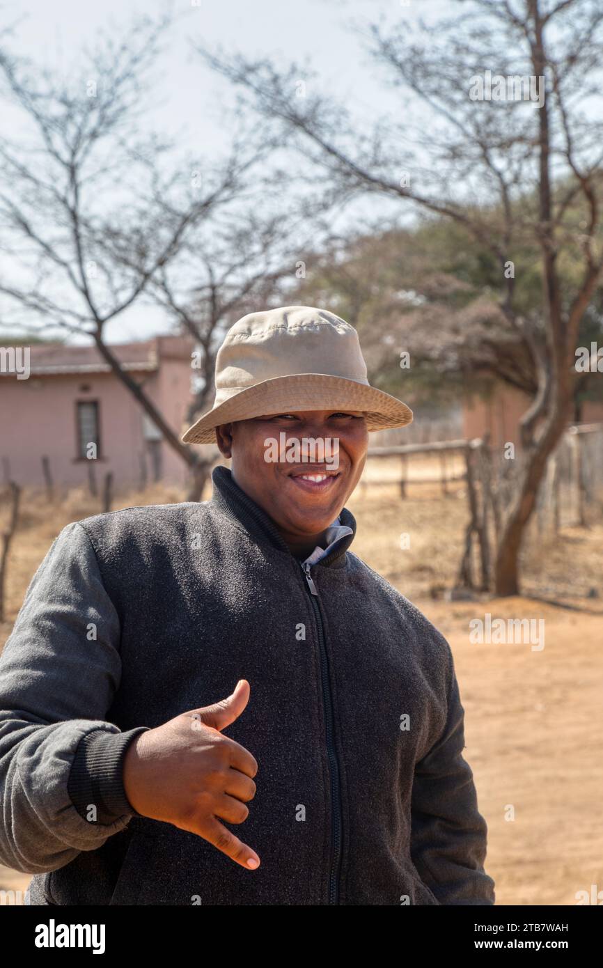 homme africain de village avec un chapeau tenant sa main avec signe ok, maison et chemin de terre en arrière-plan Banque D'Images