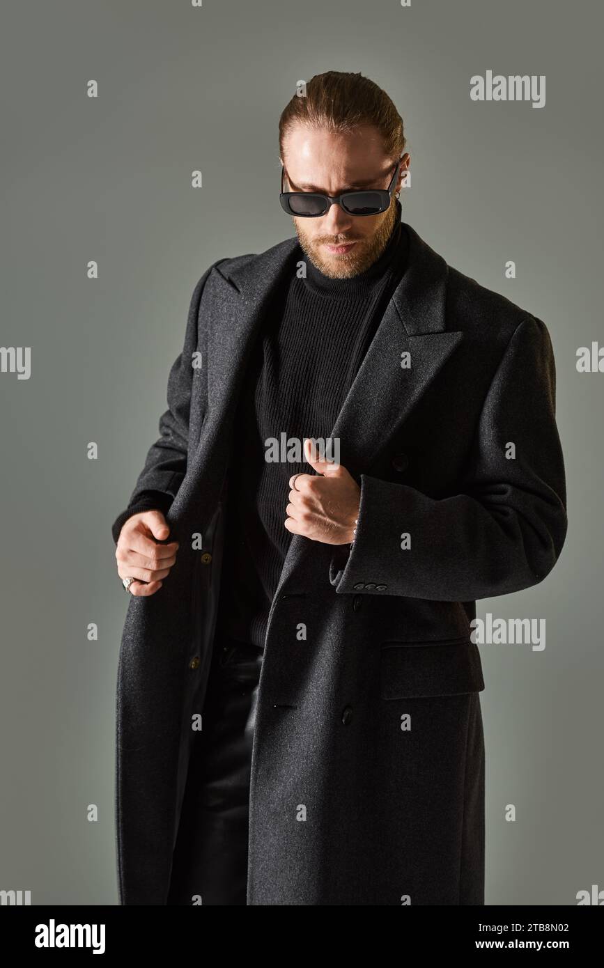 portrait de beau modèle masculin dans des lunettes de soleil à la mode et tenue noire debout sur fond gris Banque D'Images