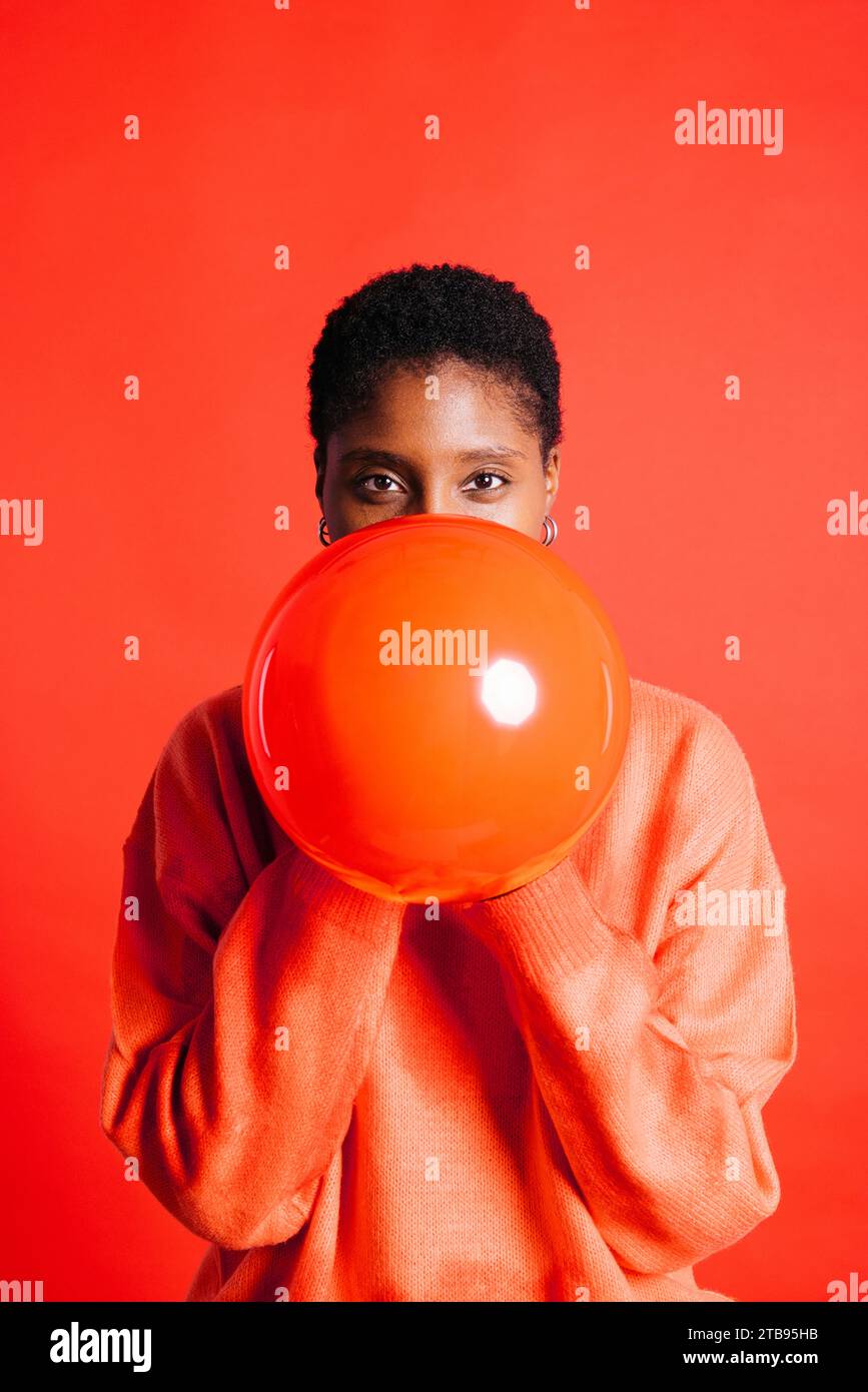 Portrait d'une jeune femme heureuse soufflant un ballon rouge devant un fond rouge dans un studio. Femme aux cheveux courts gonflant un ballon rouge. Banque D'Images Portrait d'une jeune femme heureuse soufflant un ballon rouge devant un fond rouge dans un studio. Femme aux cheveux courts gonflant un ballon rouge. Banque D'Images