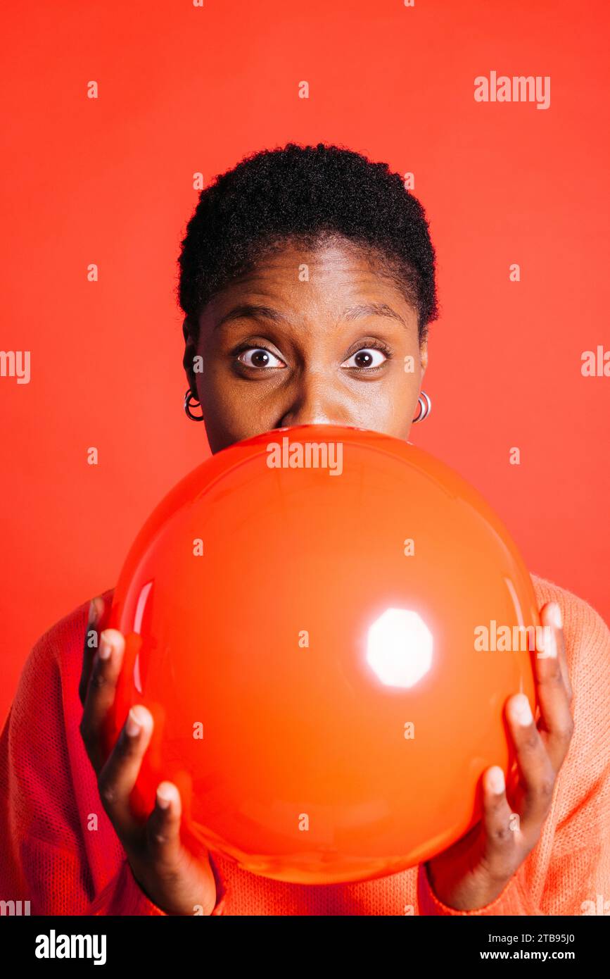 Portrait d'une jeune femme heureuse soufflant un ballon rouge devant un fond rouge dans un studio. Femme aux cheveux courts gonflant un ballon rouge. Banque D'Images Portrait d'une jeune femme heureuse soufflant un ballon rouge devant un fond rouge dans un studio. Femme aux cheveux courts gonflant un ballon rouge. Banque D'Images
