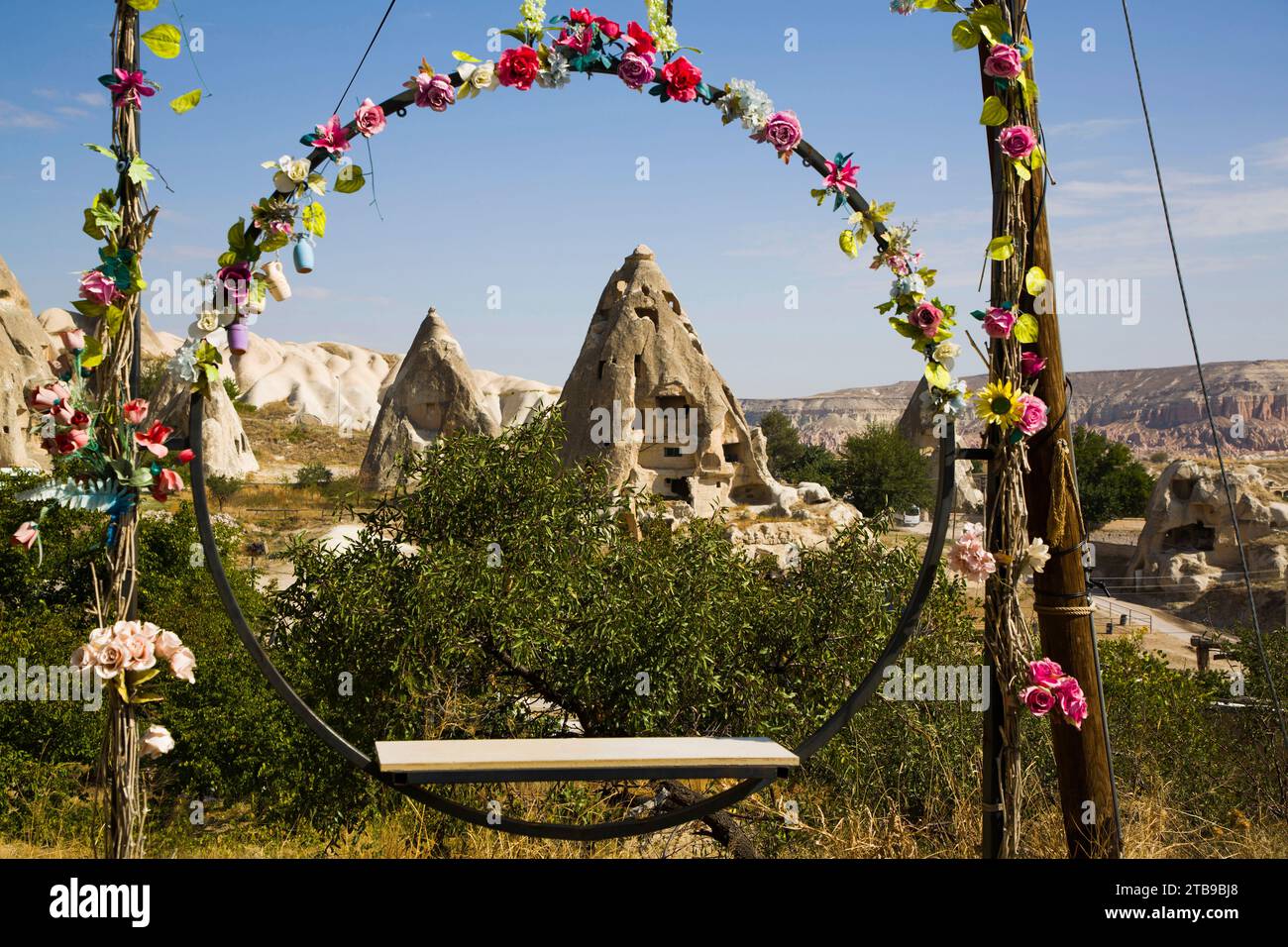 Vue à travers un siège balançoire circulaire décoré de fleurs de soie, des maisons de grotte sculptées dans les formations rocheuses contre un ciel bleu près de la ville... Banque D'Images