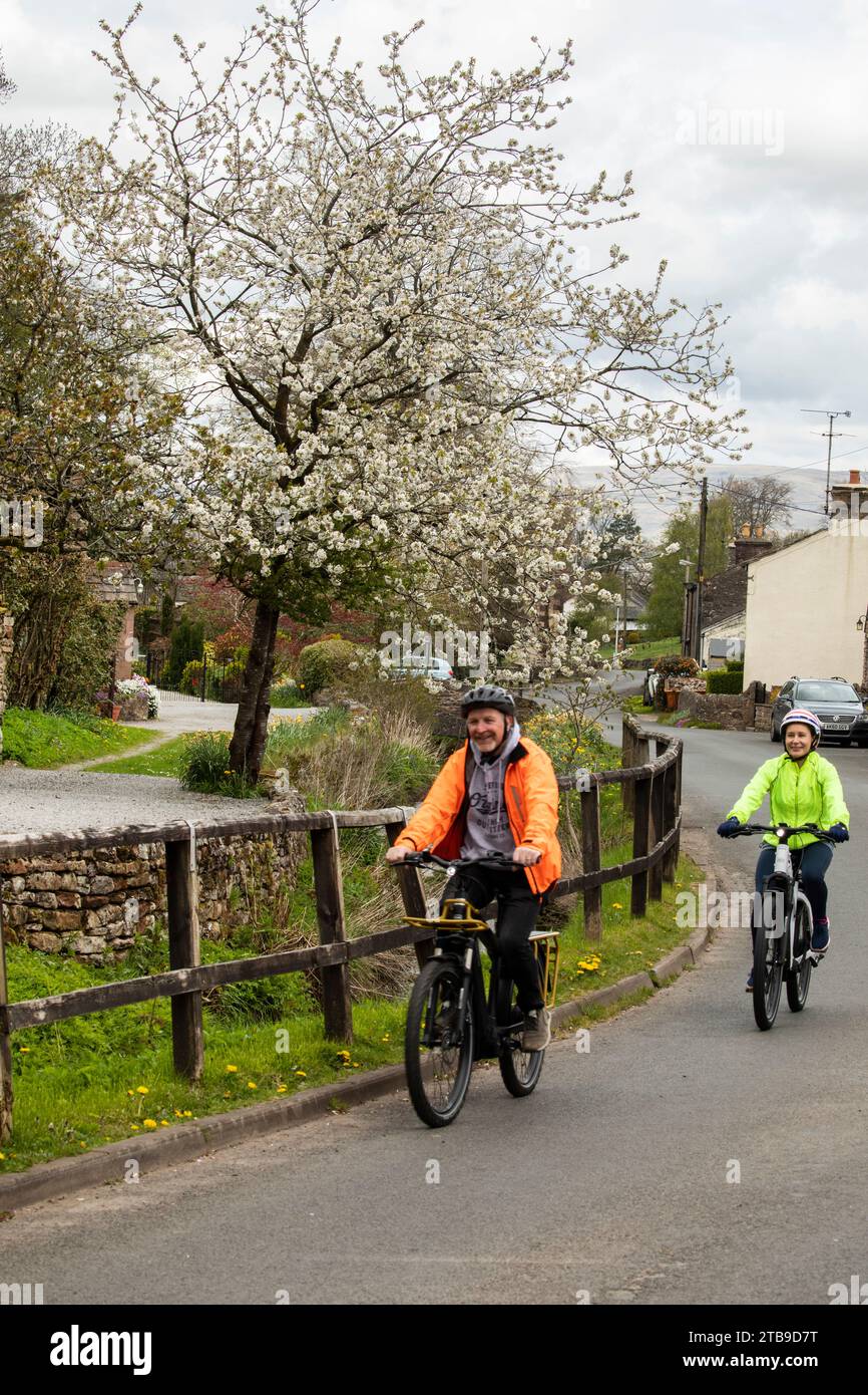 Homme et femme cyclistes à vélo électrique dans un joli village au printemps, Hartley. Kirkby Stephen, North Pennines, Cumbria Banque D'Images