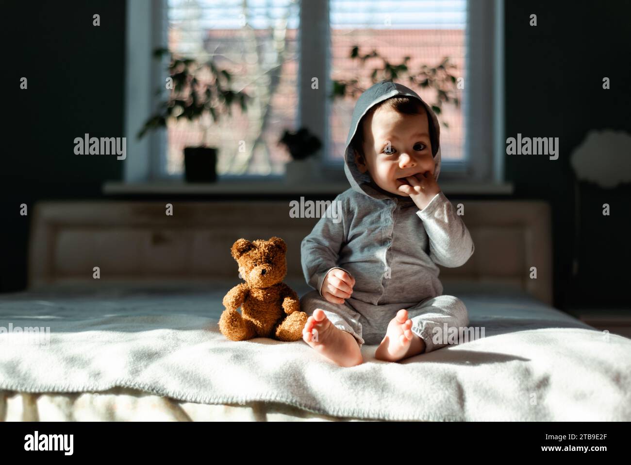 Petit garçon en pyjama gris assis sur le lit avec son ours en peluche jouet préféré. Concept d'enfance heureuse Banque D'Images