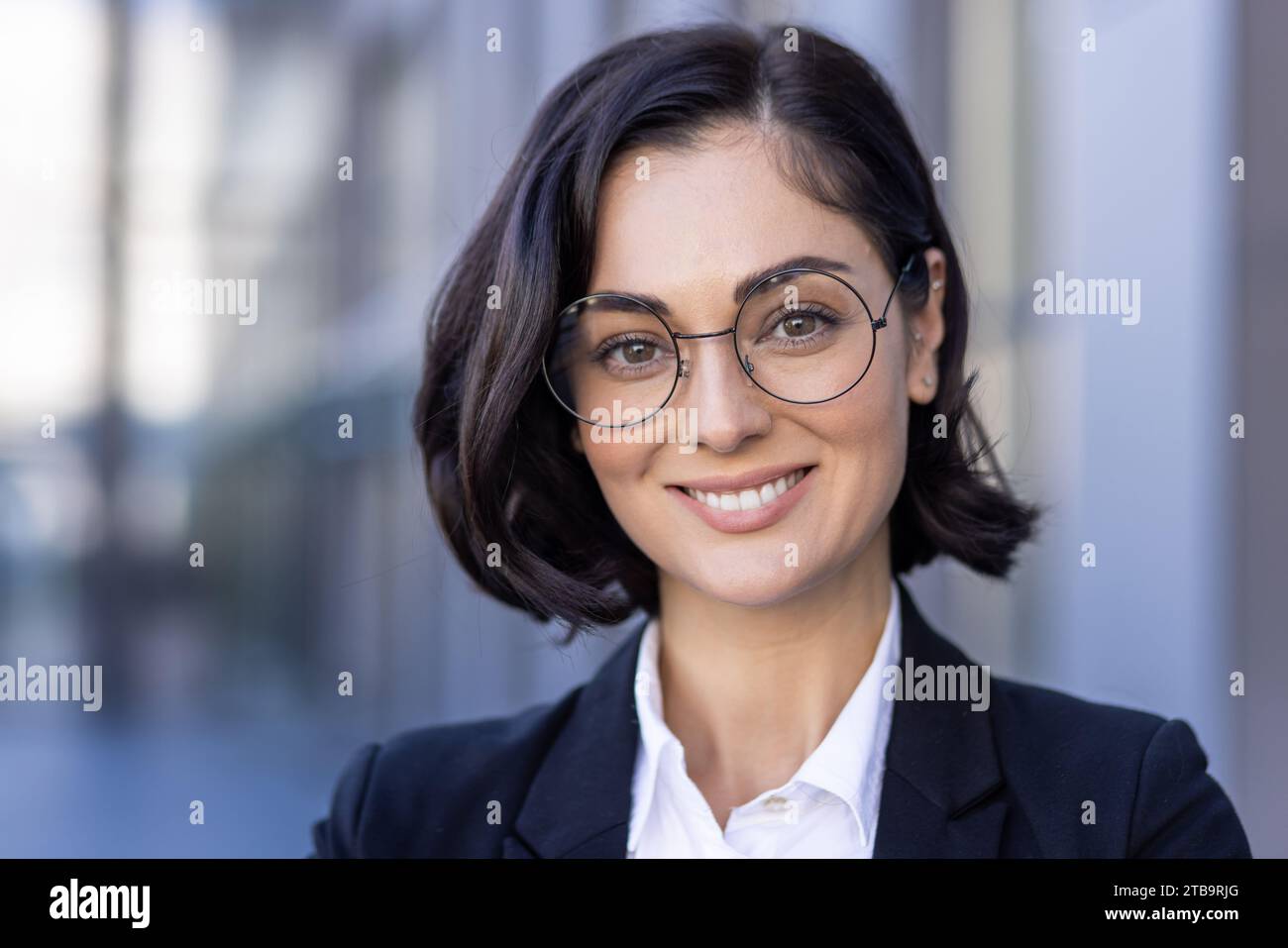Photo en gros plan d'une jeune belle avocate réussie dans un costume et des lunettes souriant à la caméra et debout à l'extérieur. Banque D'Images