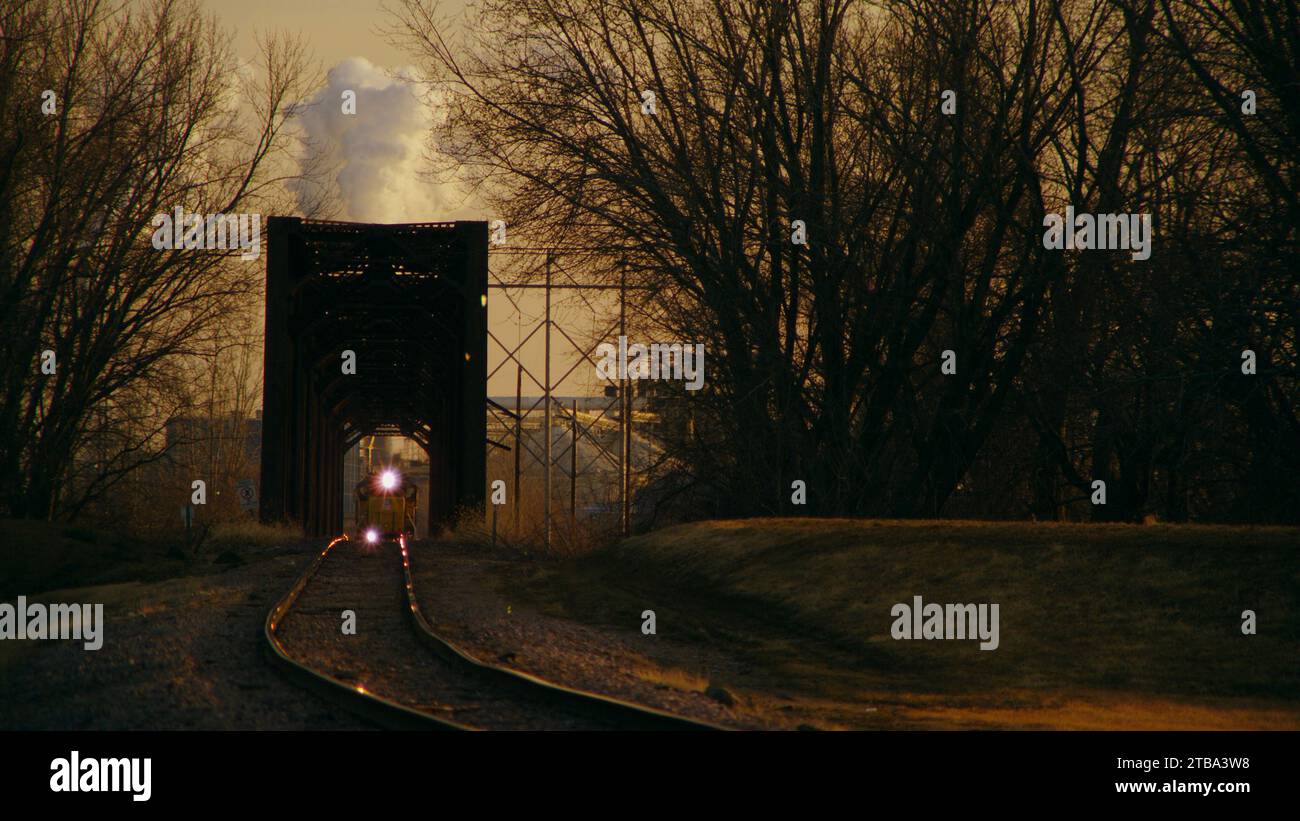 Train de marchandises partant de la centrale industrielle de combustion de charbon avec cheminée de vapeur créent une illusion d'optique lorsque la locomotive traverse un pont en treillis de fer. Banque D'Images