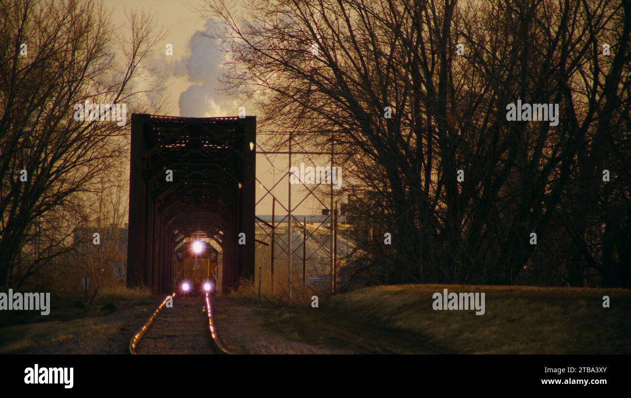 Train de marchandises quittant une centrale industrielle de combustion de charbon avec cheminée de vapeur créant une illusion d'optique lorsque la locomotive traverse un pont en treillis de fer. Banque D'Images