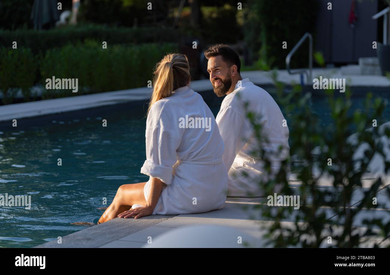 Couple en robes blanches assis dos à dos au bord d'une piscine dans un hôtel spa Banque D'Images