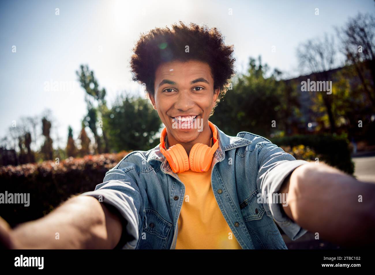 Portrait de beau mec joyeux avec chevelure porter des écouteurs de chemise en denim élégant blog faisant selfie souriant dans le parc à l'extérieur Banque D'Images