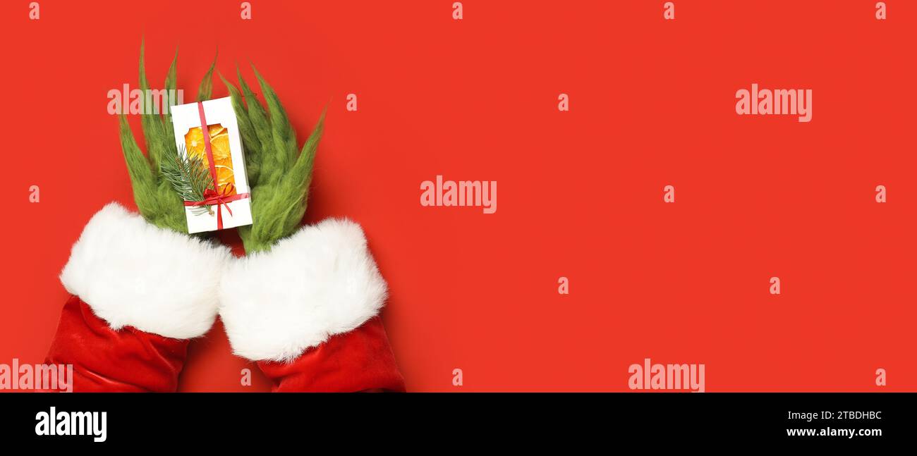 Mains velues vertes de créature dans le costume de Père Noël tenant cadeau de Noël sur fond rouge avec espace pour le texte, vue de dessus Banque D'Images