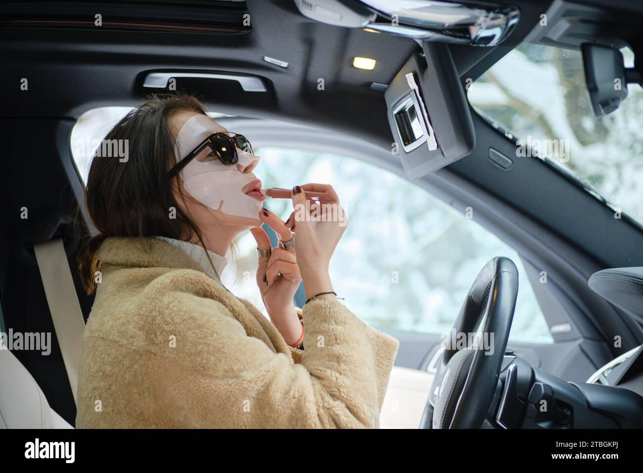 Concept du rythme rapide de la vie - femme appliquant du rouge à lèvres portant un masque facial tout en conduisant une voiture Banque D'Images